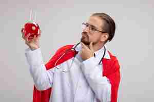 Free photo thinking young superhero guy wearing medical robe with stethoscope and glasses holding and looking at chemistry glass bottle filled with red liquid putting hand on chin isolated on white background