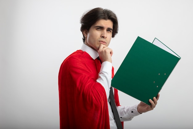 Thinking young superhero guy wearing tie holding clipboard and grabbed chin isolated on white background