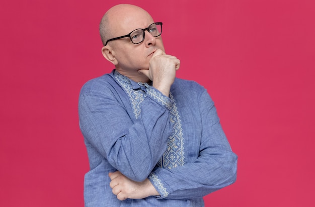 Thoughtful adult man in blue shirt wearing glasses holding his chin and looking at side 