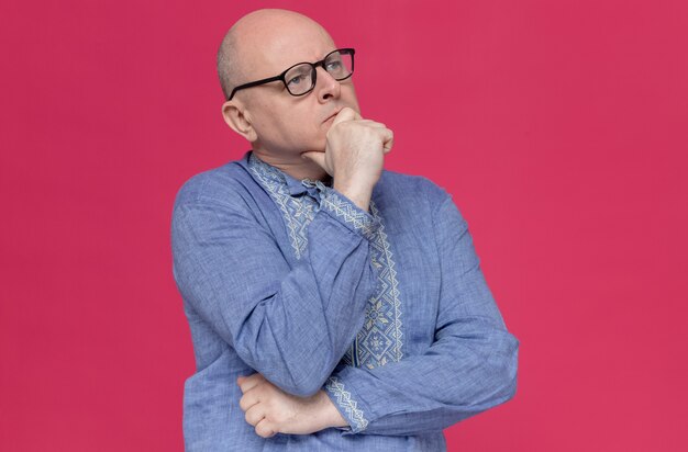 Thoughtful adult man in blue shirt wearing glasses holding his chin and looking at side 