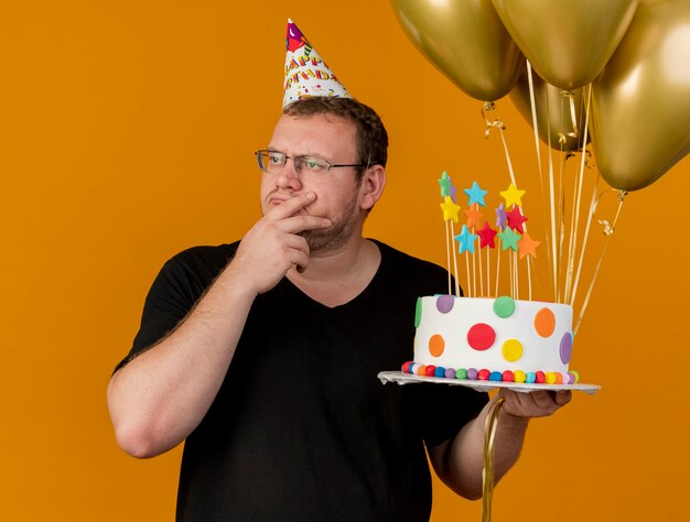 Thoughtful adult slavic man in optical glasses wearing birthday cap puts hand on chin holds helium balloons and birthday cake looking at side 