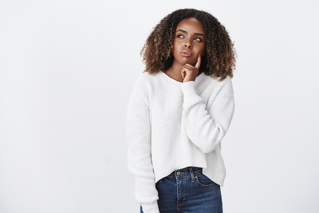 Thoughtful attractive young african american woman with curly hair in sweater deciding what wear standing over white wall squinting as thinking, touching cheek