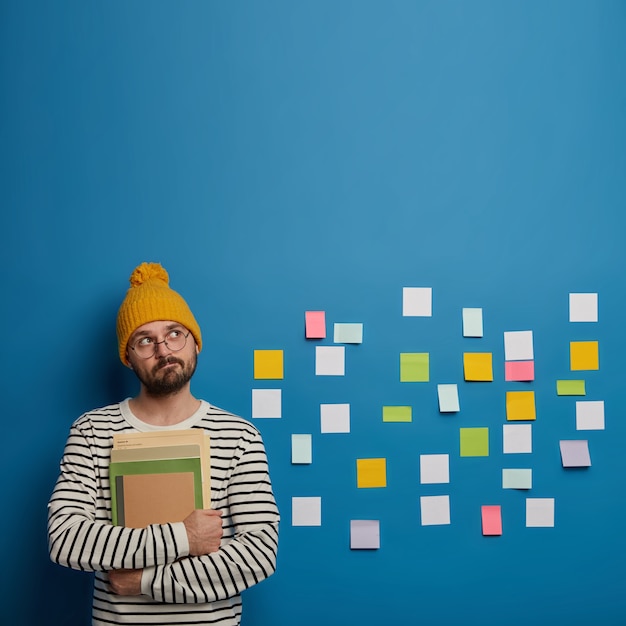 Free photo thoughtful bearded guy holds books, looks pensively above, thinks about how to make project, brainstorms different ideas in mind