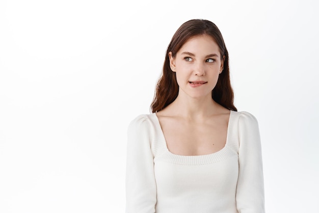 Free photo thoughtful beautiful girl wants taste something tasty biting lip from temptation looking aside at intriguing thing standing pensive against white background