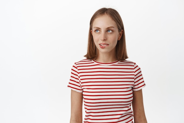 Thoughtful blond woman wants something, biting lip and looking aside with tempted look, has desire, wants to try something tasty, standing against white wall.