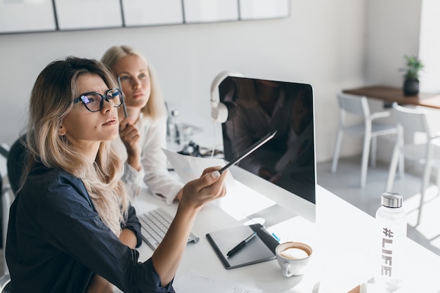 Free photo thoughtful blonde woman in glasses holding pencil and looking away during work in office. indoor portrait of busy long-haired female accountant using computer.