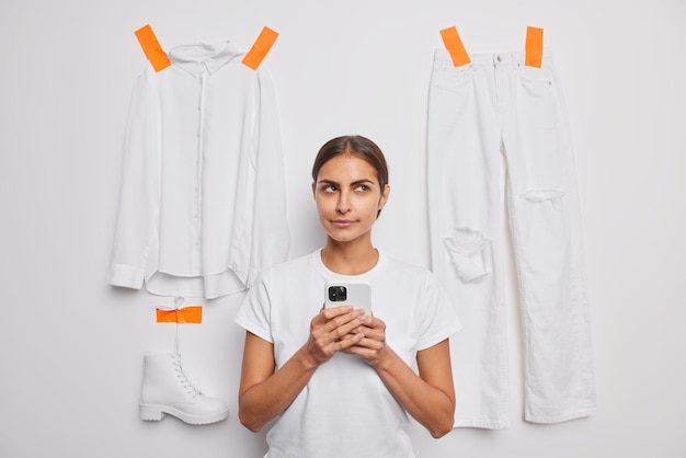 Free Photo thoughtful brunette woman dressed in casual t shirt uses mobile phone for making shopping online sells items of clothes in internet poses against white background. people and clothing concept