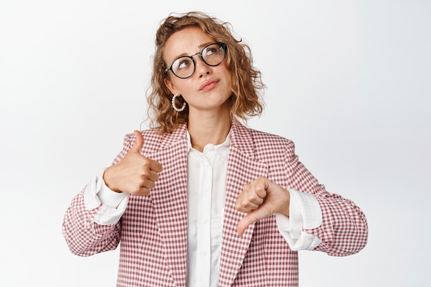 Thoughtful businesswoman in glasses and suit, showing thumbs up and down, thinking about something, making decision, weighing pros and cons on white.