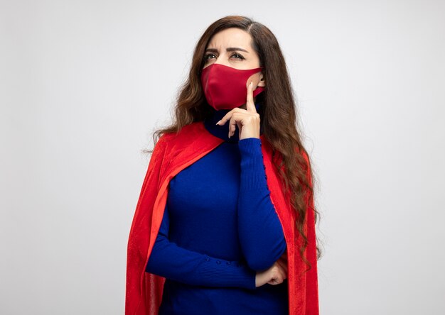 Thoughtful caucasian superhero girl with red cape wearing red protective mask puts hand on chin looking at side isolated on white wall with copy space