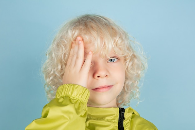 Thoughtful. Close up portrait of beautiful caucasian little boy isolated on blue  wall. Blonde curly male model. Concept of facial expression, human emotions, childhood, 