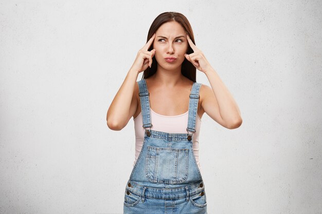 Thoughtful concentrated female in denim overalls, keeping fingers on temples, trying to get idea, looking pensively aside, isolated over white blank wall. Young woman trying recall right answer