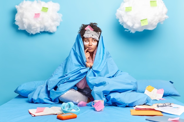 Free photo thoughtful dark skinned woman looks aside tries to decide something wrapped in soft blanket poses on bed writes ideas on stickers and notebook