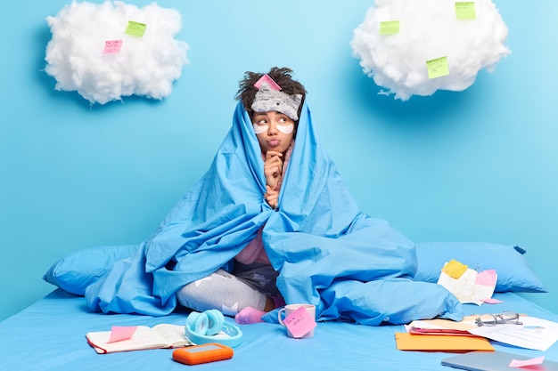 Free photo thoughtful dark skinned woman wrapped in soft blue duvet keeps lips rounded is thinking.