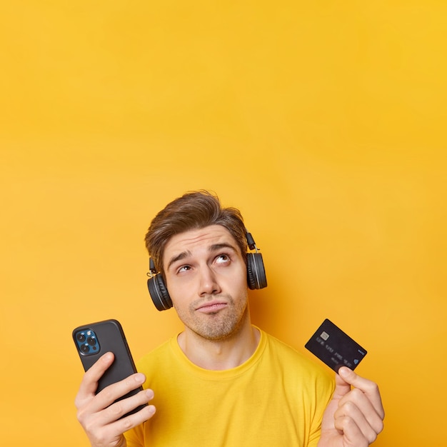 Thoughtful displeased man concentrated above listens music via headphones holds cellphone and credit card thinks how to make payment on website isolated over yellow wall blank space overhead