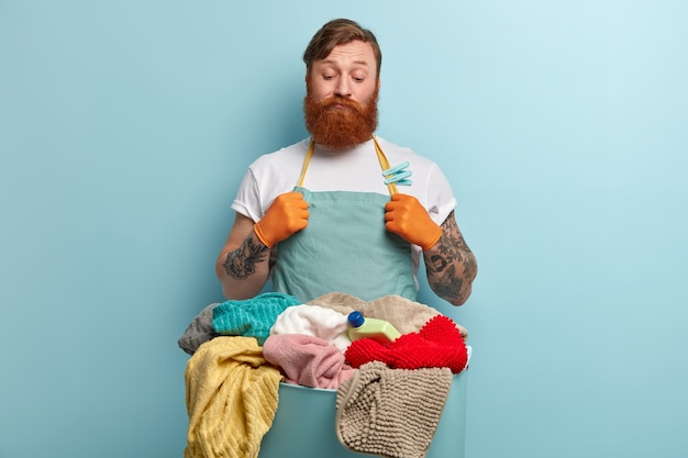 Free Photo thoughtful doubtful indecisive man with thick ginger beard, looks at laundry, doesnt know how to wash, isnt eager to do washing