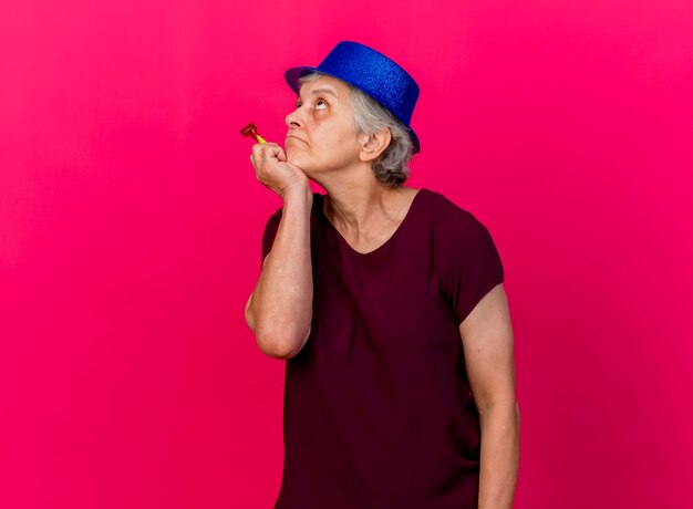 Thoughtful elderly woman wearing party hat puts hand on chin holding whistle looking up on pink