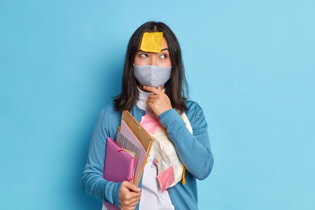 Thoughtful female entrepreneur works from home as social distancing wears disposable mask thinks how to prepare research work wears casual jumper.