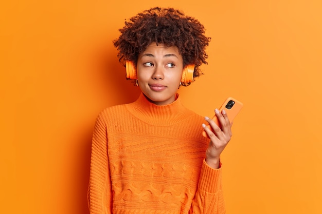 Thoughtful female meloman listens audio track from playlist holds modern smartphone looks pensively aside