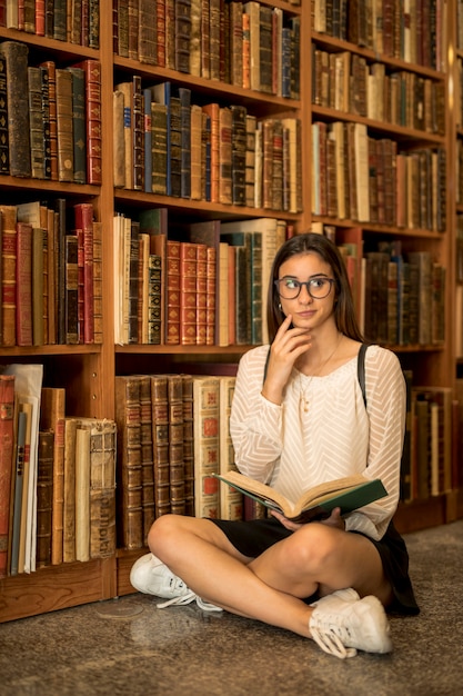 Thoughtful female student sitting cross-legged with book in library