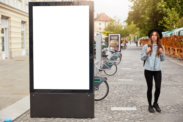 Thoughtful female tourist strolls on footway near lightbox with mock up blank space for your advertising content or commercial information. Street style concept. Focus on billboard at sidewalk