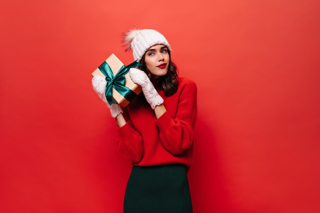 Thoughtful girl shakes gift box with green bow Young curly woman in red sweater white knitted mittens and hat poses on isolated