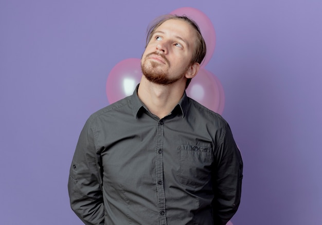 Thoughtful handsome man holds helium balloons behind and looks up isolated on purple wall