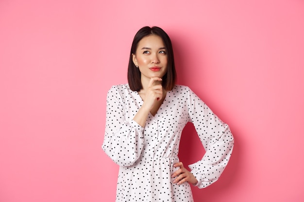 Free photo thoughtful happy asian woman making decision smiling satisfied and looking at upper left logo thinki...