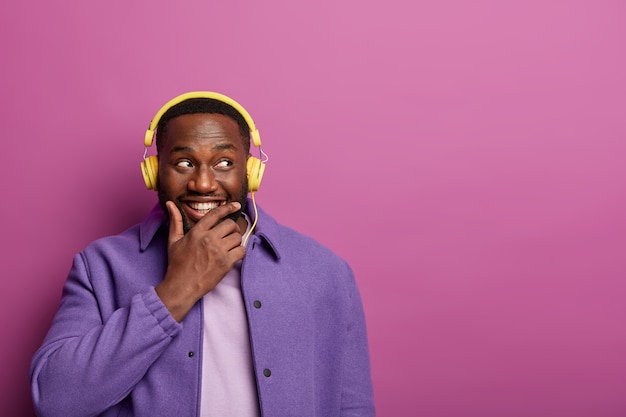 Thoughtful happy man holds chin, bought new headphones for listening music, looks aside