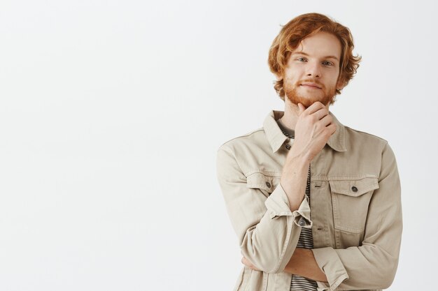 Thoughtful and intrigued bearded redhead guy posing against the white wall