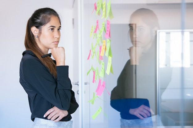 Thoughtful Latin businesswoman holding marker and reading notes on glass wall. Focused confident pretty female worker in suit thinking about idea for project. 