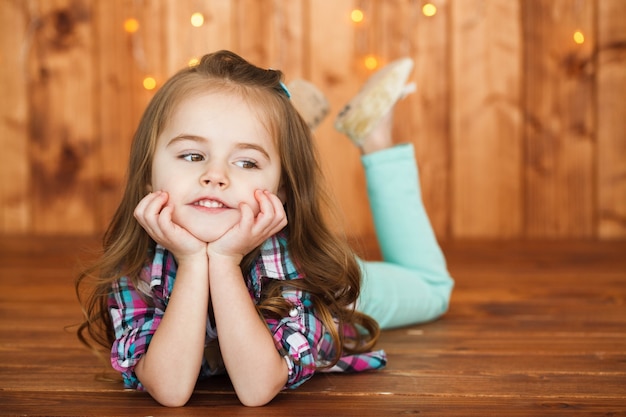 Thoughtful little girl lies on wooden floor before wall with yellow lights