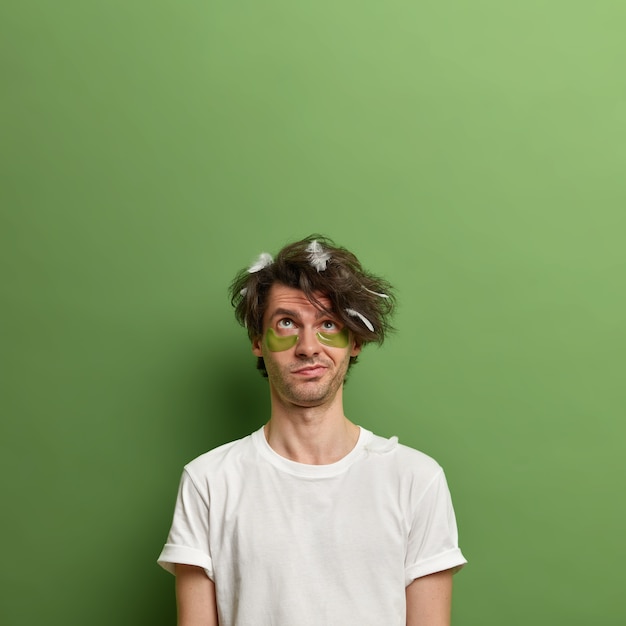 Free photo thoughtful man concentrated above, waits for nice effect after applying collagen patches under eyes, has uncombed hair with feathers, poses against green wall, copy space for your promotion