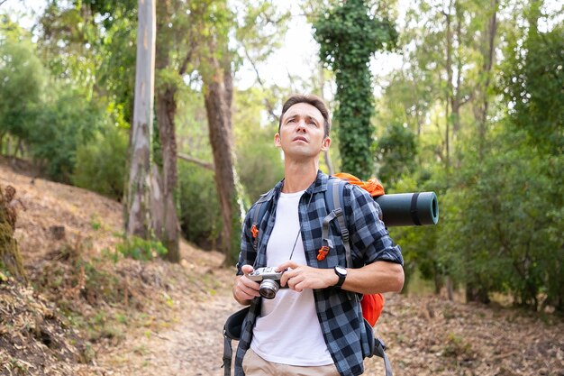 Free Photo thoughtful man holding camera, looking away and standing on road. caucasian tourist exploring nature and taking photo of nature. tourism, adventure and summer vacation concept