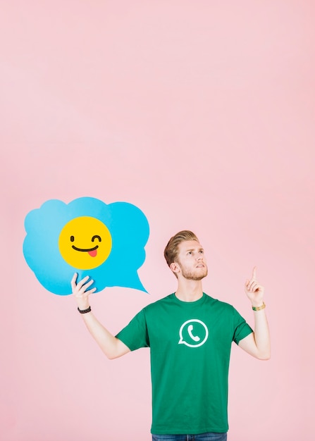 Free photo thoughtful man pointing upward while holding winking emoji speech bubble