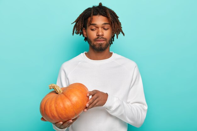 Thoughtful man with dreads, holds pumpkin, prepares for Halloween, wears white sweater, has beard