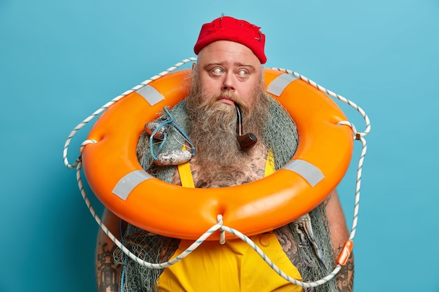 Free photo thoughtful mariner carries inflated ring buoy on neck, ready to rescue people at sea, has thick long beard, smokes pipe