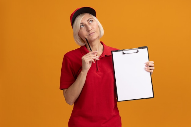 Free Photo thoughtful middle-aged blonde delivery woman in red uniform and cap holding clipboard touching face with pencil looking up isolated on orange wall with copy space