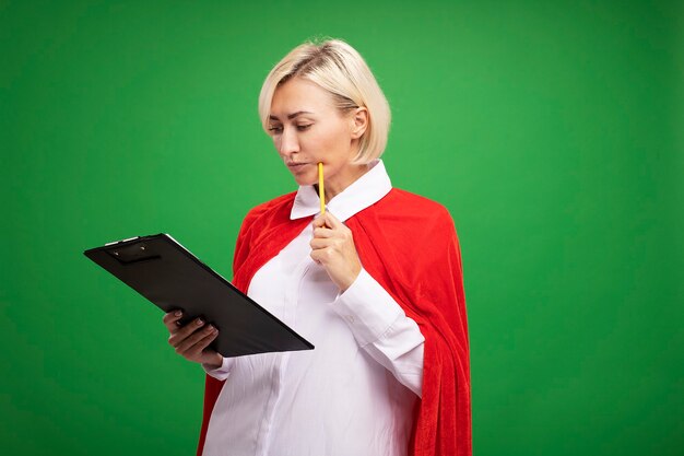 Thoughtful middle-aged blonde superhero woman in red cape holding and looking at clipboard touching cheek with pencil 