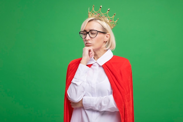 Free photo thoughtful middle-aged blonde superhero woman in red cape wearing glasses and crown putting hand on chin looking down isolated on green wall with copy space