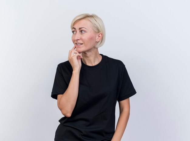 Thoughtful middle-aged blonde woman touching chin looking at side isolated on white wall