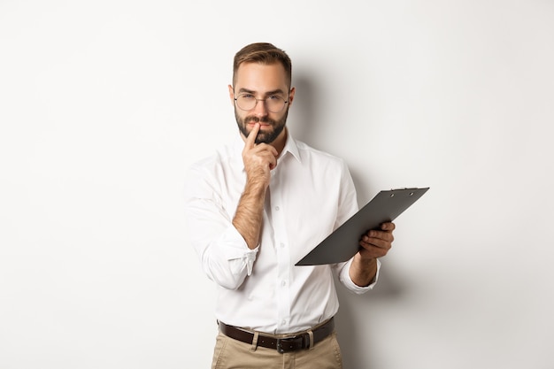 Thoughtful office employee working, holding documents on clipboard and thinking, standing  