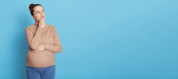 Thoughtful pregnant woman looks thoughtfully aside, keeping hand on chin, makes planning about child birth, dreams to become mother, wearing beige sweater and jeans, isolated on blue wall.