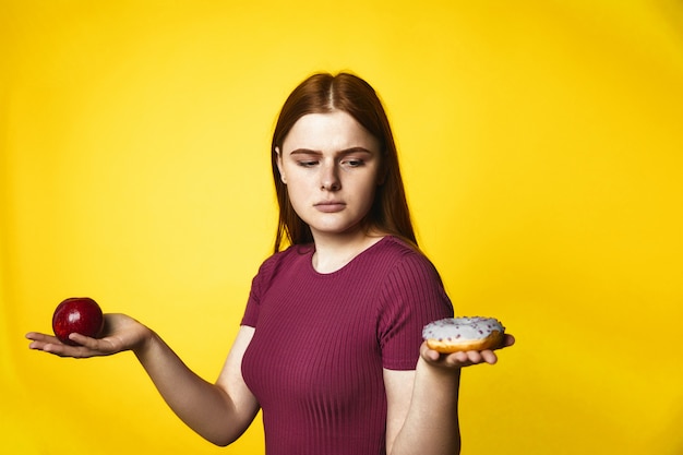 Thoughtful redhead caucasian girl is holding apple in one hand and donut in another