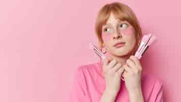 Free photo thoughtful redhead teenage girl with freckled skin undergoes beauty treatments applies hydrogel patches under eyes holds cosmetic brushes near face poses against pink background blank space