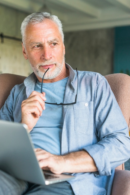 Thoughtful senior man holding eyeglasses sitting in armchair with laptop