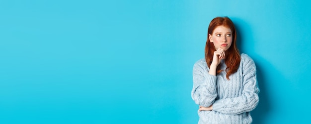 Free photo thoughtful and upset redhead girl looking right pondering solution standing in sweater against blue