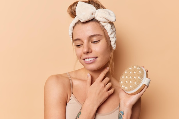 Free photo thoughtful woman concentrated down touches face gently holds dry brush for exfoliating skin wears headband and t shirt isolated over beige background. cellulite treatment and dry brushing concept