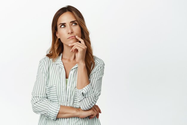 Thoughtful woman, wife thinking, looking up and touch lip, ponder important decision, making choice, standing in blouse against white wall