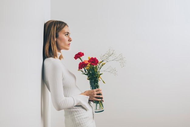 Free photo thoughtful woman with flowers in vase at wall