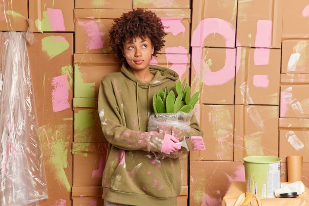 Thoughtful young Afro American woman poses alone in new apartment holds paint brush and thinks.